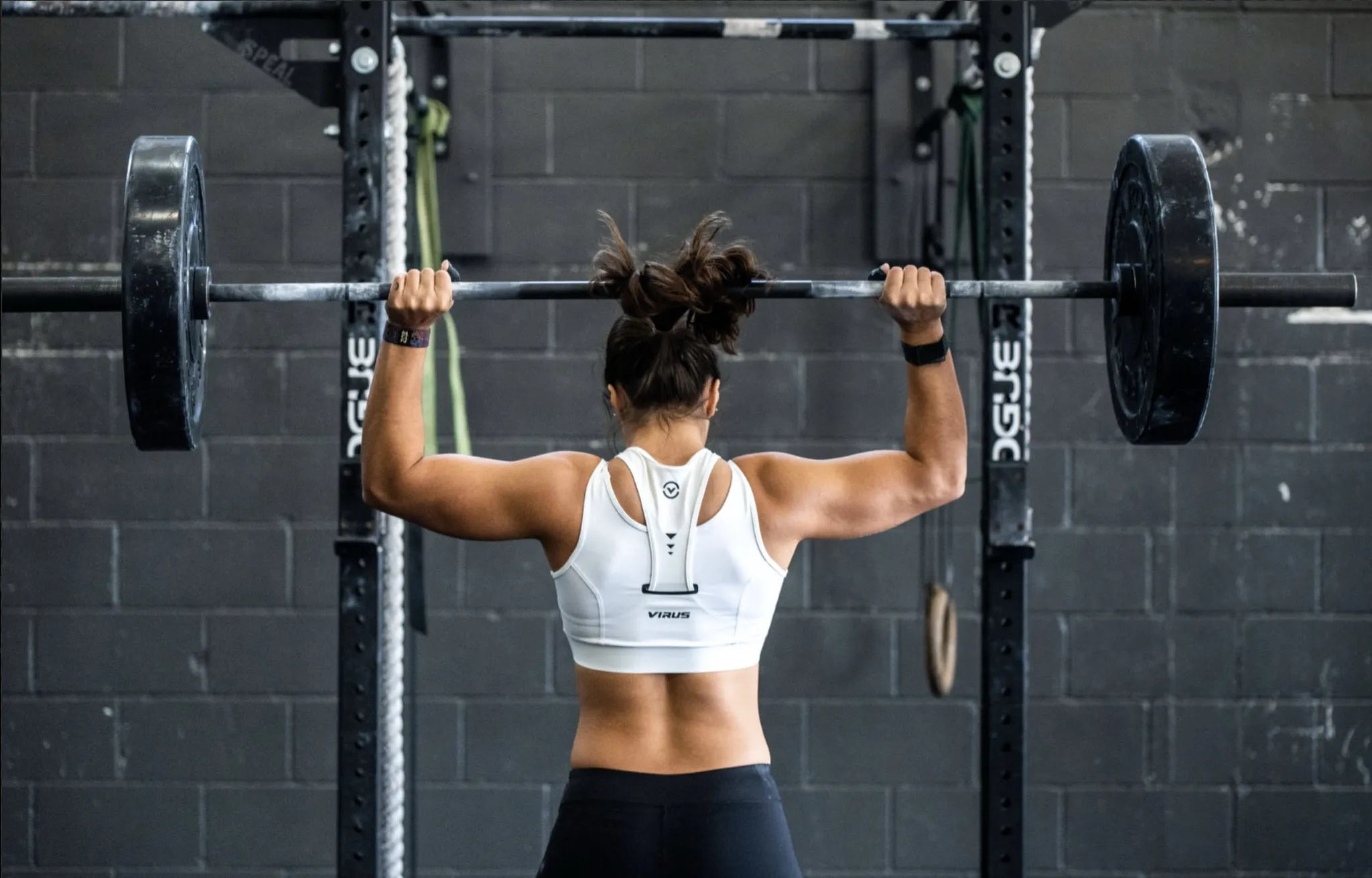 A woman in a gym presses a loaded barbell behind her neck, seen from behind